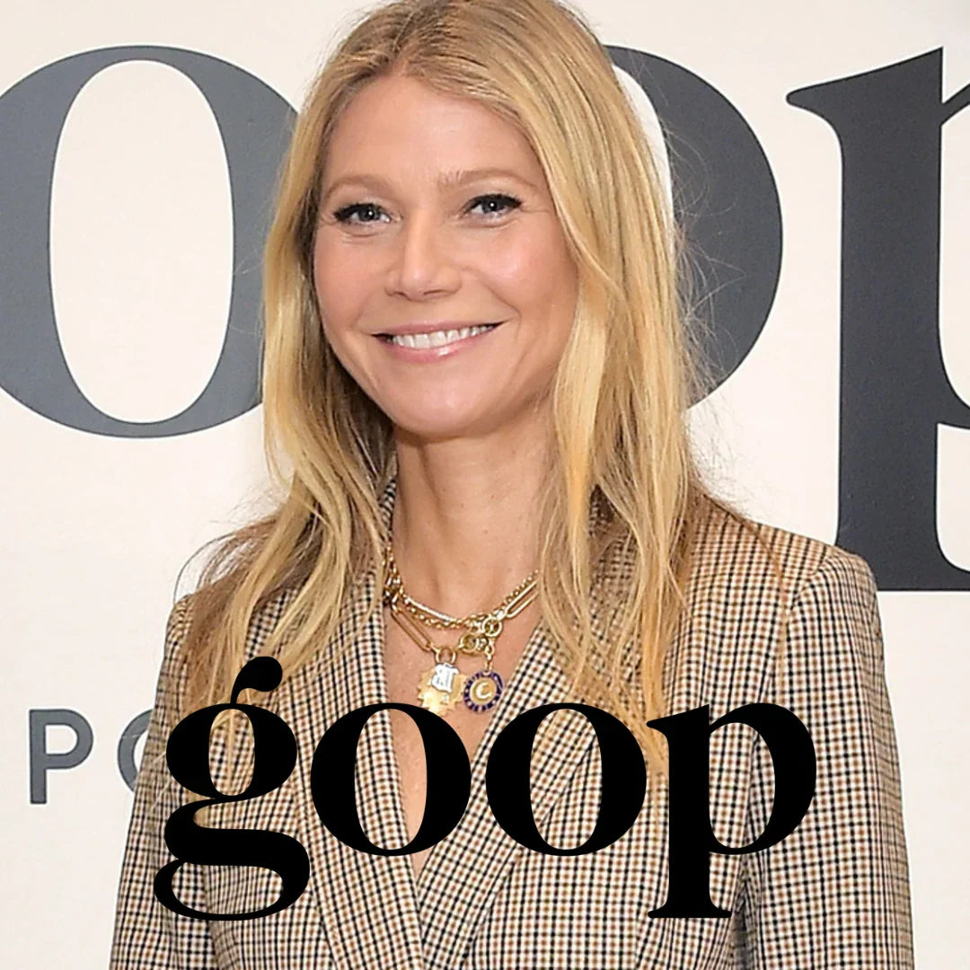 A smiling blonde woman in a beige and brown checkered blazer, wearing layered gold necklaces, standing in front of a white event backdrop with large dark lettering. A large black 'goop' logo watermark is overlaid across the bottom.

