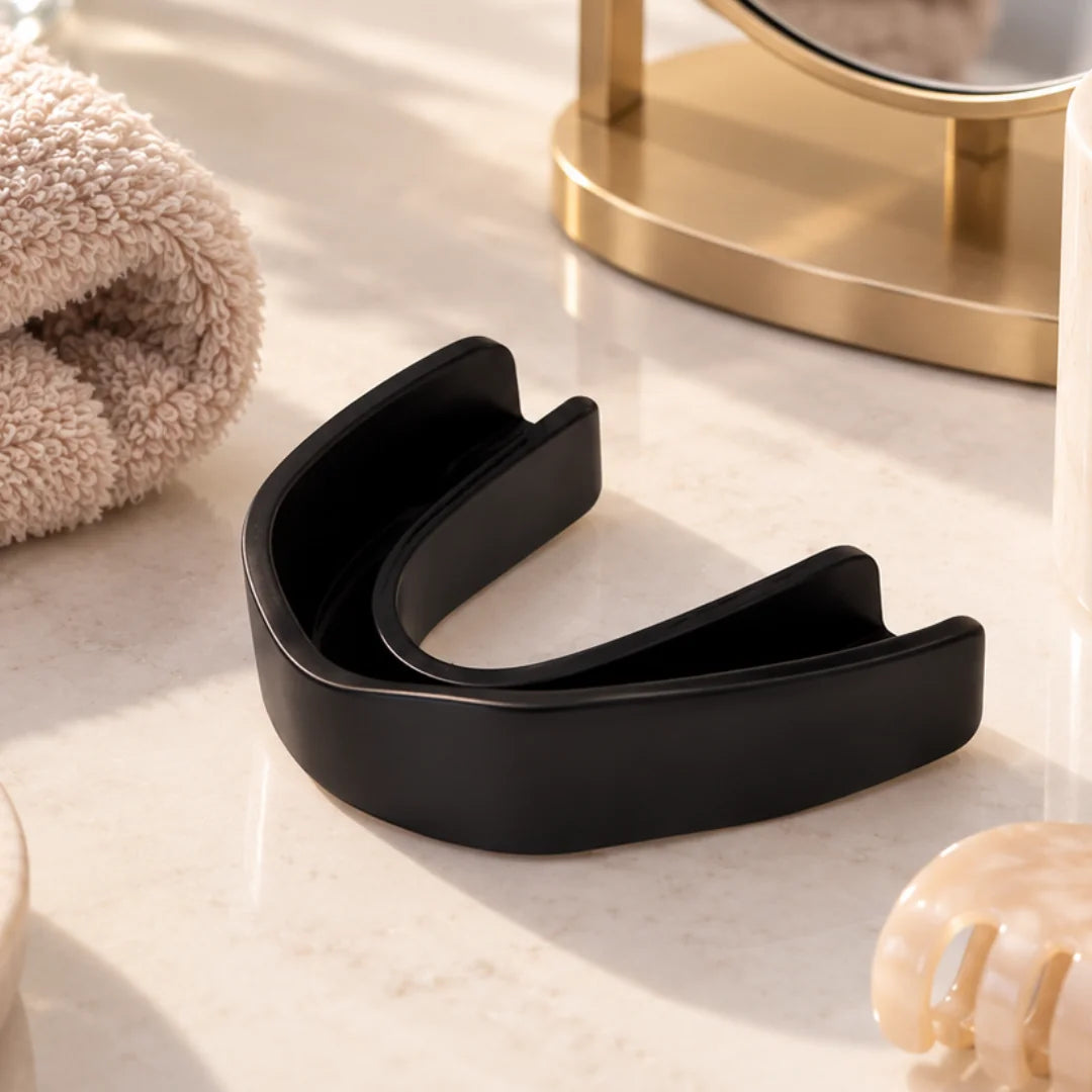 Black facial aligner mouthpiece resting on a marble vanity beside a folded towel, gold mirror stand, and hair clip.